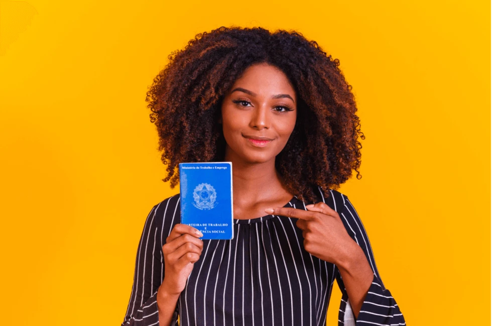 Jovem mulher afrodescendente com cabelo cacheado segurando a carteira de trabalho com fundo amarelo vibrante.
