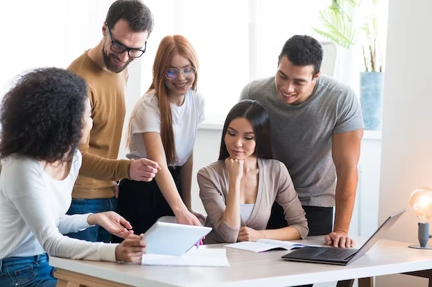 Grupo de cinco jovens profissionais colaborando em reunião de equipe em um escritório, compartilhando ideias e usando dispositivos tecnológicos.