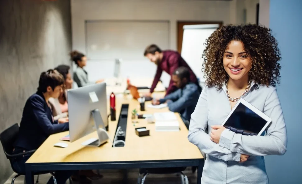 Empresária jovem e confiante em reunião de negócios com colegas, usando roupas formais e sorrindo, em ambiente corporativo moderno e colaborativo.