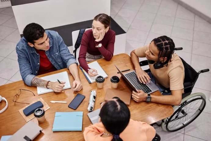 Reunião de equipe com diversidade, incluindo uma pessoa em cadeira de rodas, discutindo estratégias de trabalho colaborativo em um ambiente corporativo.
