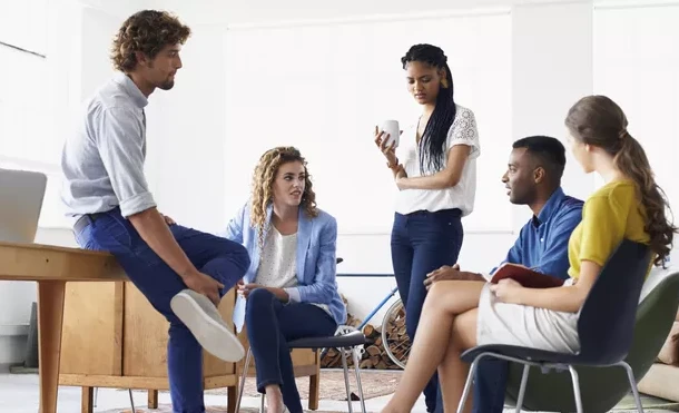 Grupo de jovens participando de uma sessão de terapia de grupo, incluindo uma mulher terapeuta anotando, enquanto os outros conversam e escutam em ambiente claro.